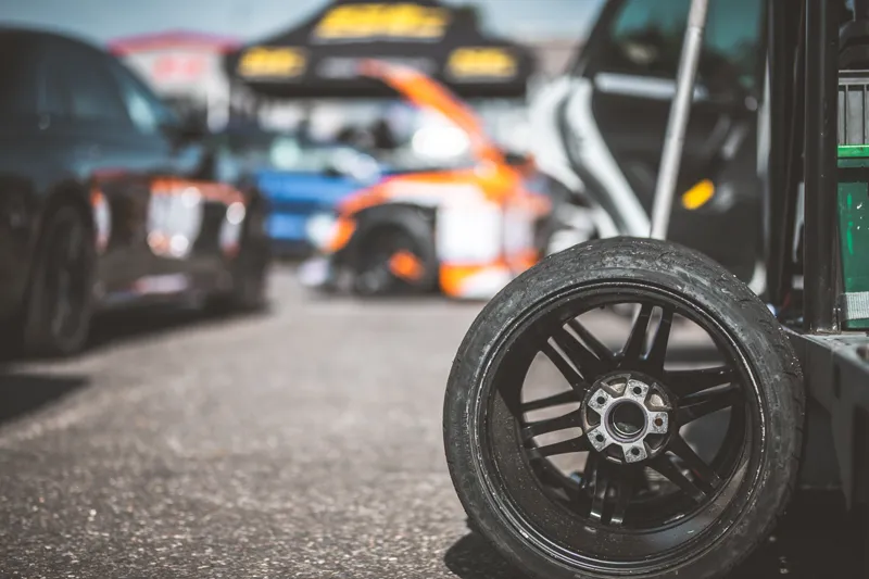 Close-up of a worn racing tire and wheel in the paddock area, with performance cars and equipment blurred in the background during a track event—highlighting the risks covered by Track Day Insurance.
