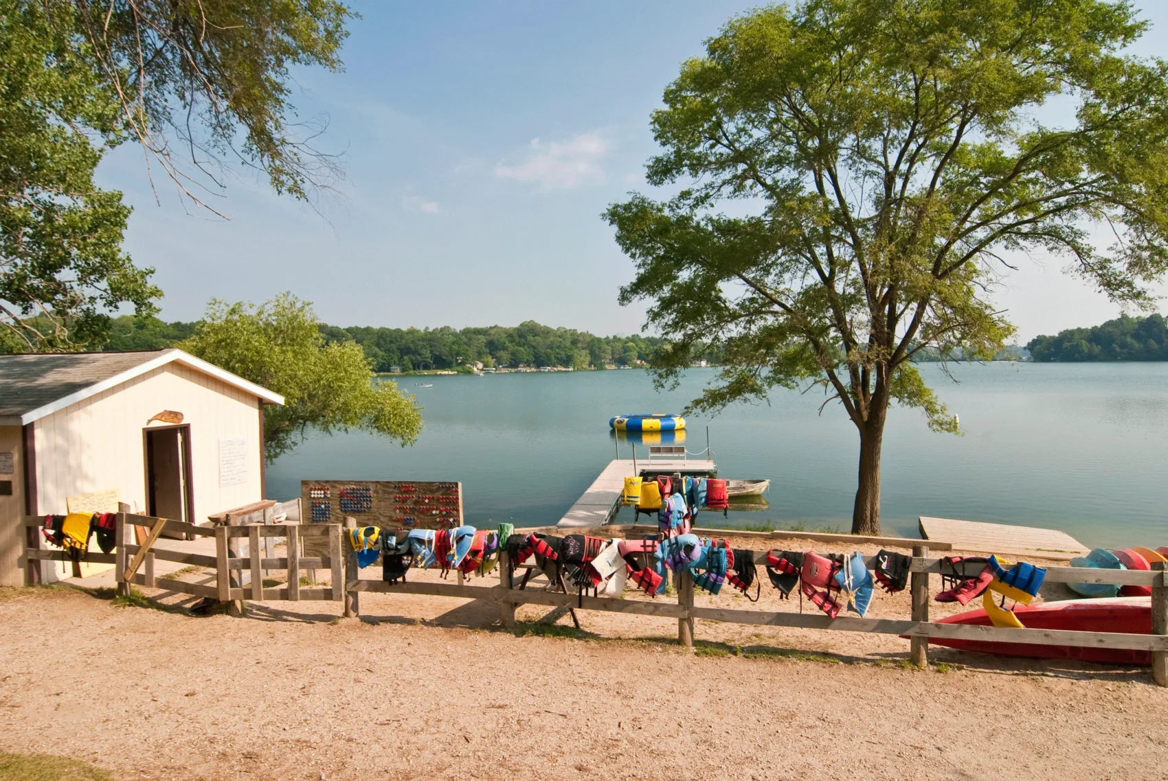 Lakeside summer camp waterfront with a dock, canoes, and colorful life jackets hanging on a fence beside a calm lake.