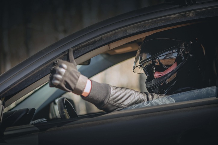 Driver wearing a black helmet and gloves giving a thumbs-up from inside a car.