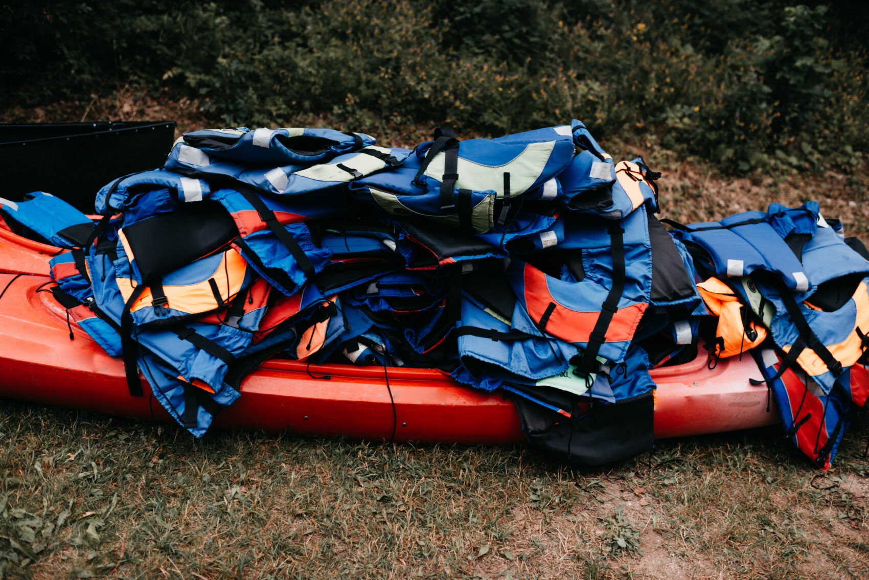 A pile of colorful life jackets stacked on a red kayak on the grass at a camp waterfront area
